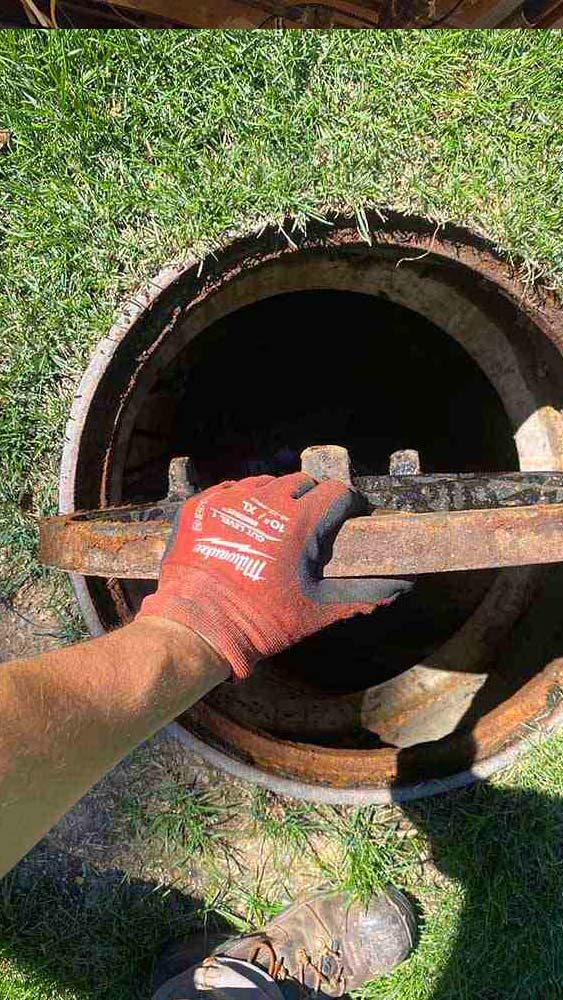 Hand in a red glove lifting a rusty sewer manhole cover on a grassy area, illustrating sewer line maintenance by Jeffrey Burke Plumbing, Heating & Cooling.